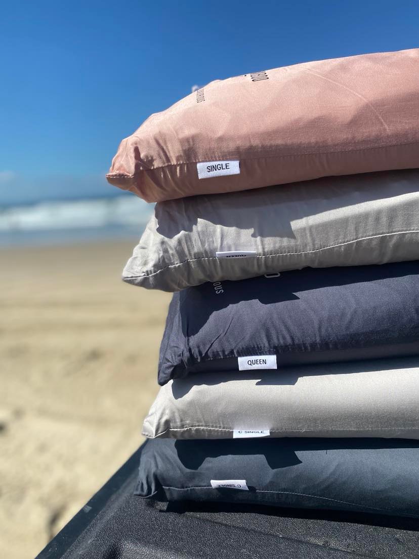Stack of organic-cotton pillows in terracotta, grey and charcoal on a beach with sea behind