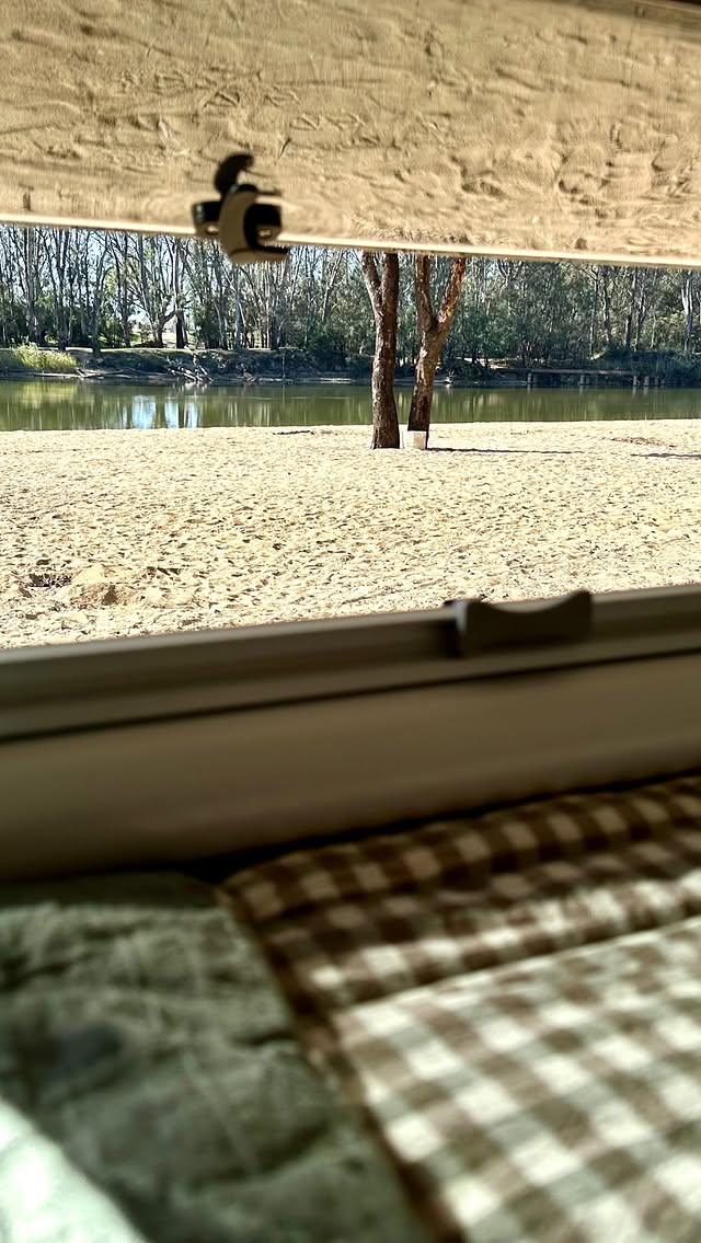 View through a caravan window onto a riverbank lined with red gums, gingham pillow in the foreground