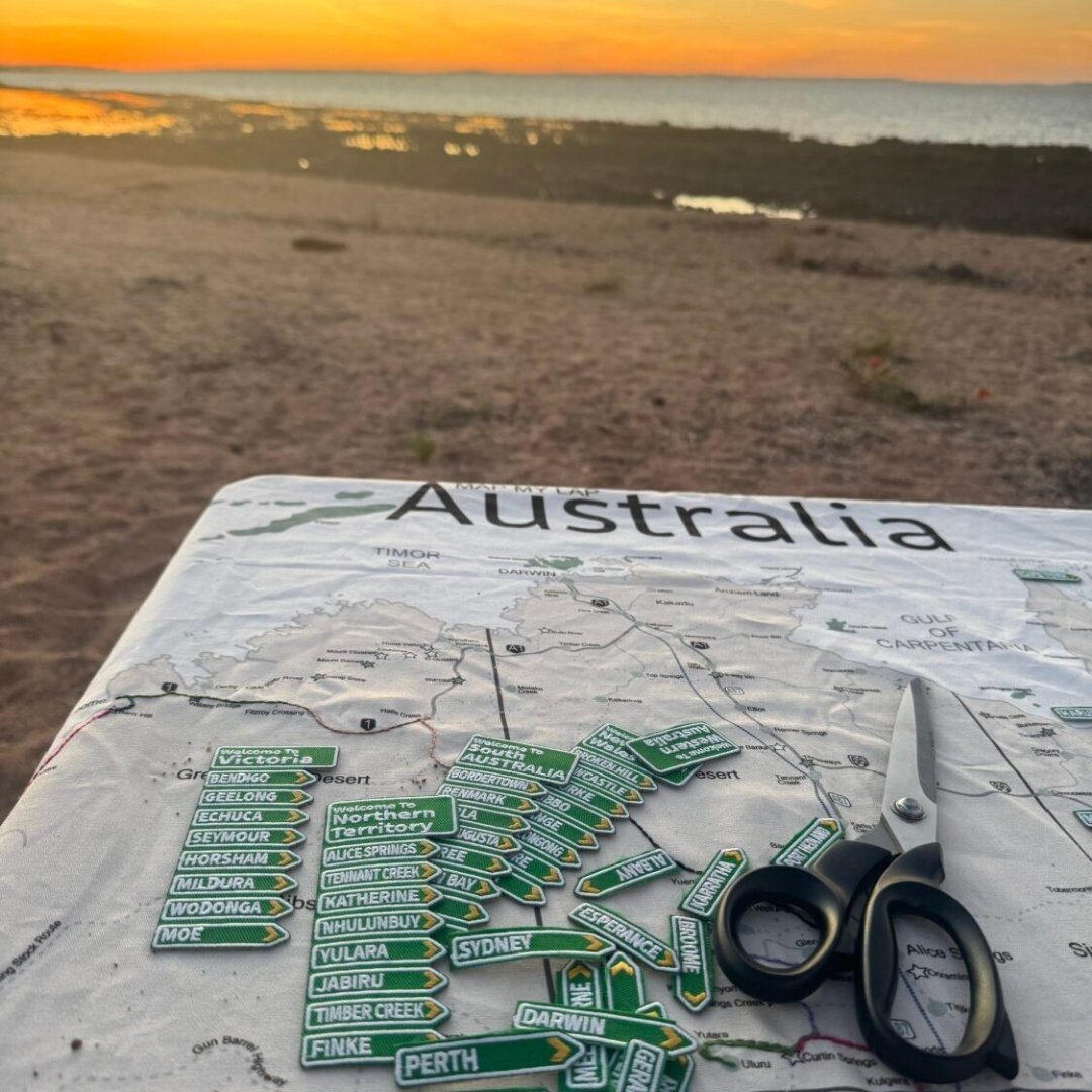 A printed Australia map fabric with embroidered town patches at sunset, scissors and threads on the sand