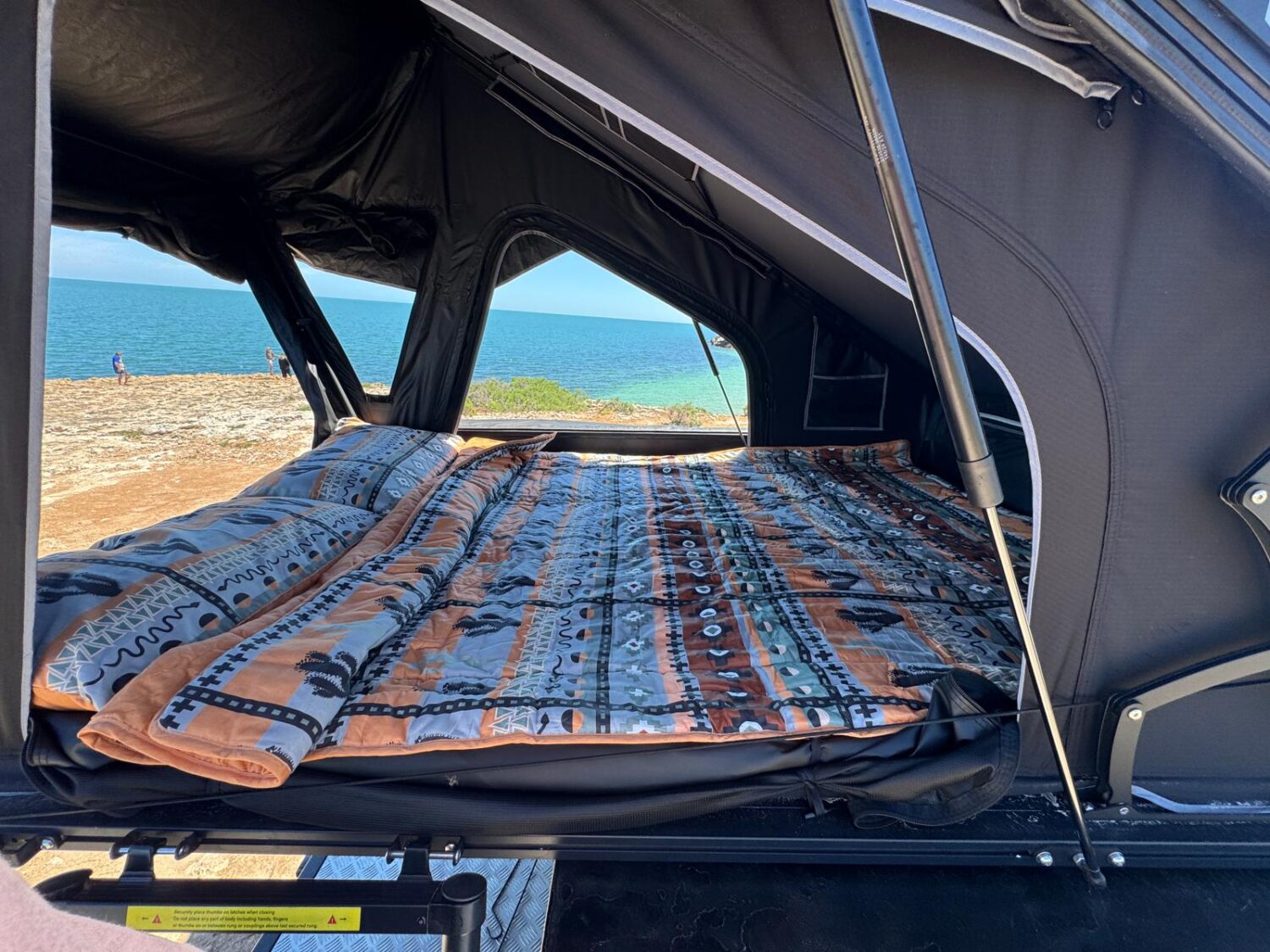 A roof top tent set up at a coastal lookout with a printed Zipluxe bed set, ocean stretching out beyond